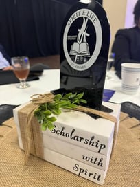 a decorative table centerpiece from an event, featuring a black, tombstone-shaped plaque with the logo and text "Spirit & Life Seminary" and "Scholarship with Spirit." The plaque is placed on top of a stack of three white, book-like blocks tied together with twine and decorated with a small green plant. The text "Scholarship with Spirit" is also written on the side of the book-like blocks. 